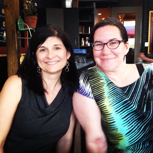 Two smiling women pose in a cafe, one in a dark cowl-neck top and one in glasses and green. — illustrating “May Favourites from Rimmell” on Florence and Mary