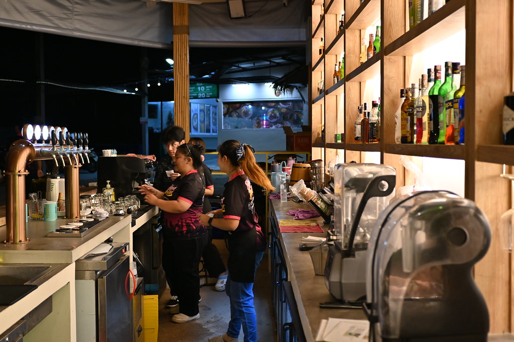 Three staff in black and pink uniforms work behind brass beer taps at a warmly lit outdoor bar. — illustrating “Autumn Glory by Pyrex” on Florence and Mary