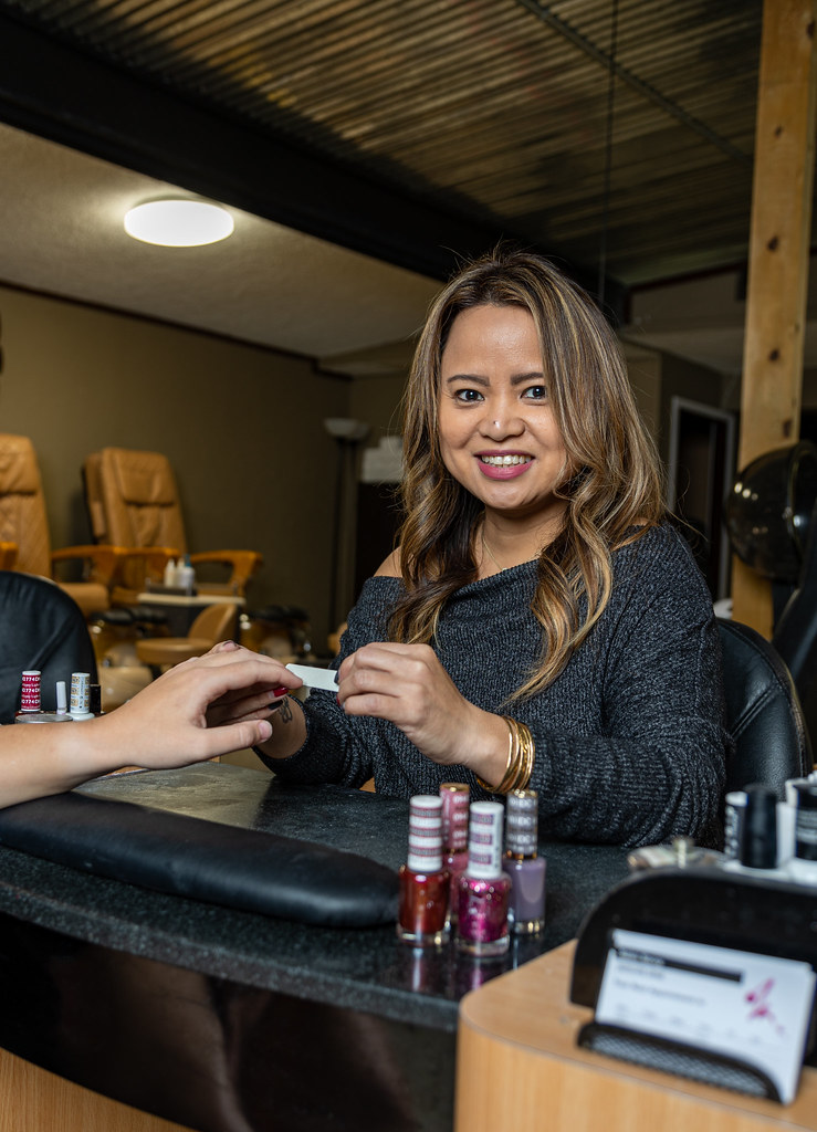 A smiling manicurist in a dark grey sweater files a customer nails beside several polish bottles. — illustrating “Barry M Confetti Nails - Sour Apple” on Florence and Mary