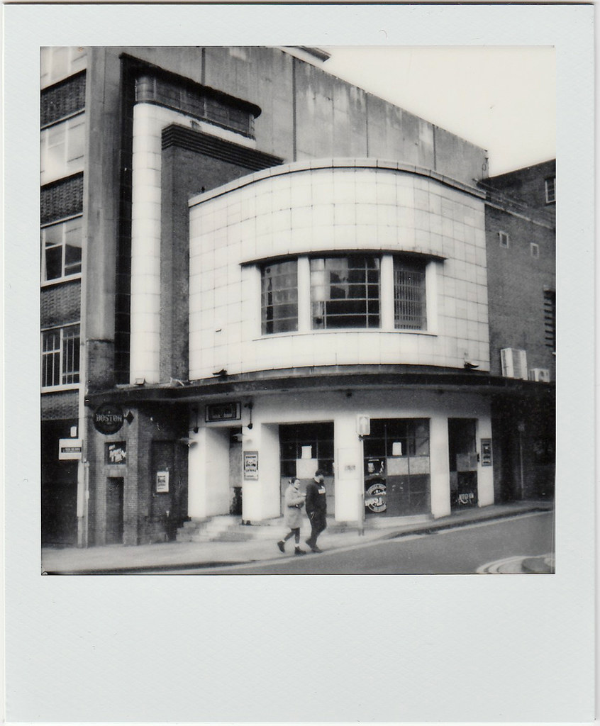 A black-and-white photo shows two people walking past a curved tiled Art Deco building on a slope. — illustrating “BEAUTY EMPTIES | April 2015” on Florence and Mary