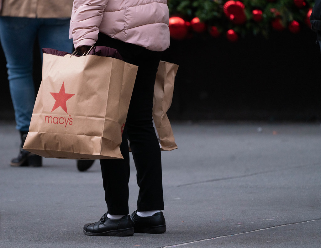 A person in a light pink puffer jacket holds a brown Macy shopping bag on a sidewalk with holiday ornaments. — illustrating “Beauty Haul and Repurchasing” on Florence and Mary