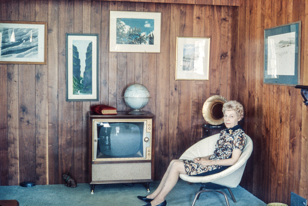 An elderly woman sits in a white swivel chair in a wood-paneled room with a vintage console television. — illustrating “Beauty Tutorials on Apple TV” on Florence and Mary