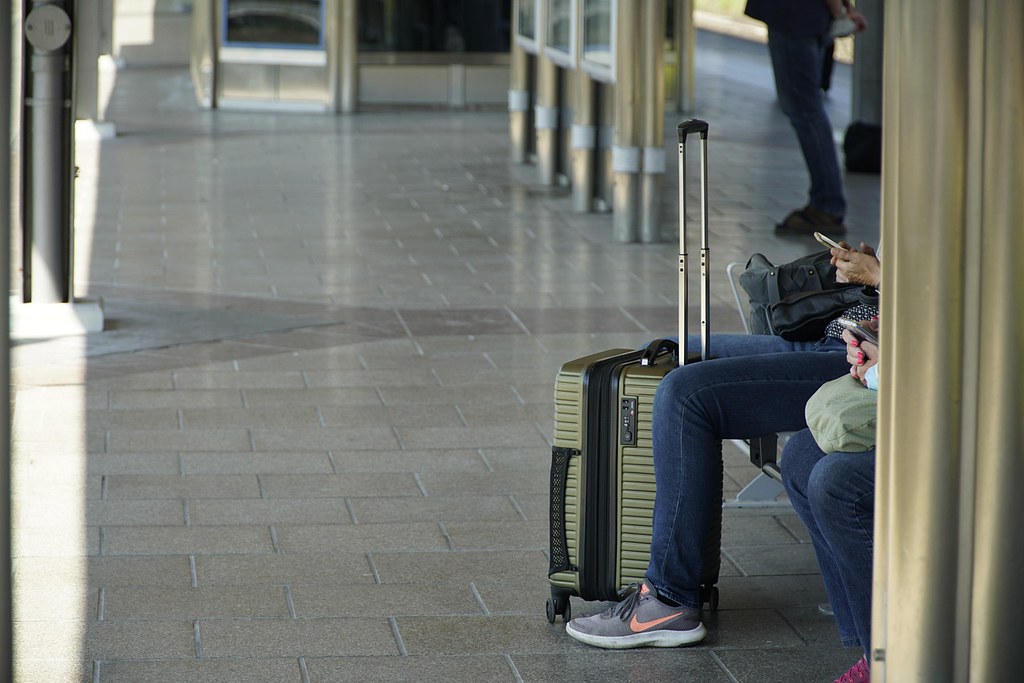 Travelers wait in a tiled transport hub beside an olive-green hardshell suitcase and gray Nike sneakers. — illustrating “BEDM: Bank Holiday Fun” on Florence and Mary