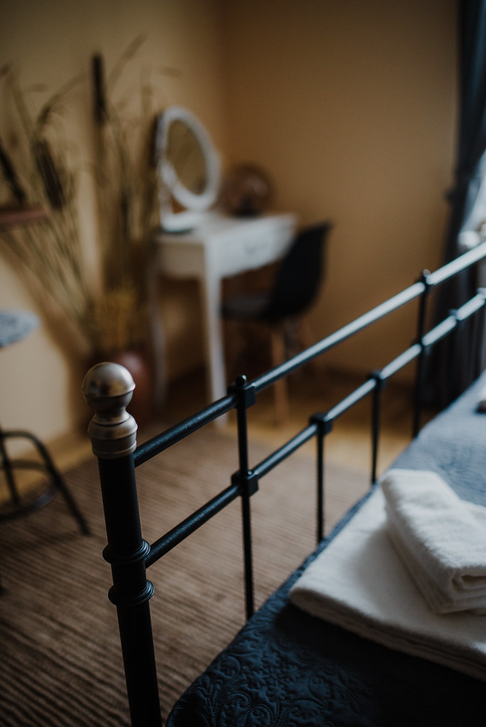 A black metal bed frame holds folded white towels on a dark bedspread with a blurred vanity behind. — illustrating “Bedroom Redesign | Somewhere in limbo” on Florence and Mary