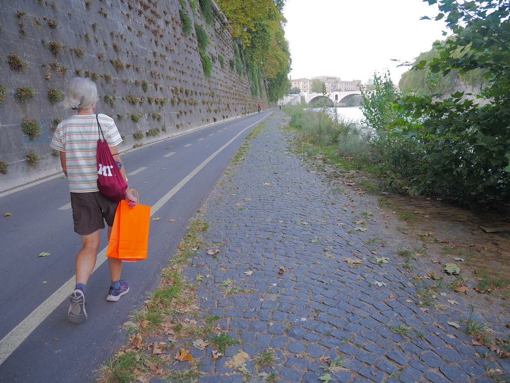 A grey-haired woman walks a riverside path carrying a bright orange shopping bag beside a high stone wall. — illustrating “Bicester Village Shopping Haul #2” on Florence and Mary