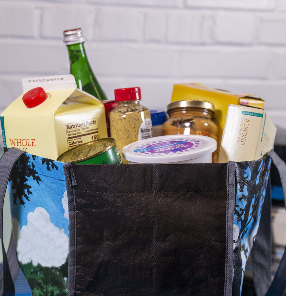 A blue and black patterned reusable bag overflows with groceries including milk and almond bars against white bricks. — illustrating “Bicester Village Shopping Haul” on Florence and Mary