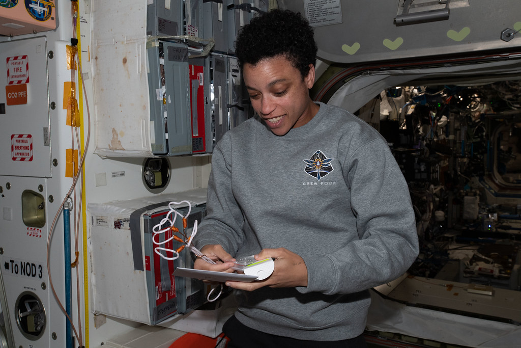 A woman in a grey Crew Four sweatshirt smiles while holding a small object inside a spacecraft storage module. — illustrating “Birthday Gifts - Part 1” on Florence and Mary