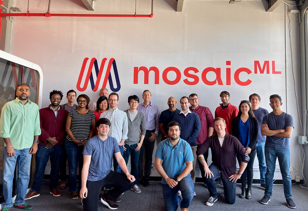 Nineteen people pose for a casual office team photo beneath a red mosaicML logo on a white wall. — illustrating “Blog Every Day in May with Rosalilium” on Florence and Mary