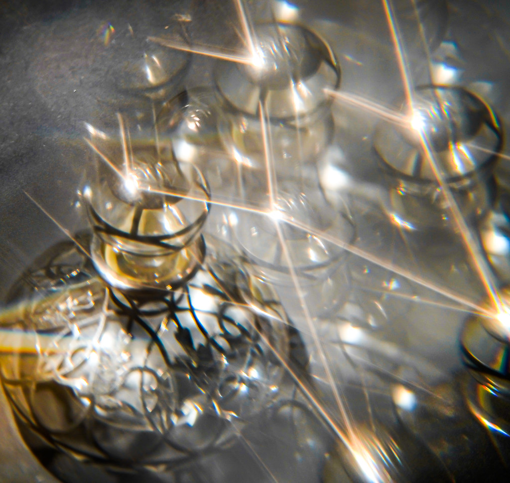 Faceted crystal decorative bottles with dark filigree patterns catch bright golden starburst light flares. — illustrating “Britney Spears Rocker Femme Fantasy Perfume” on Florence and Mary