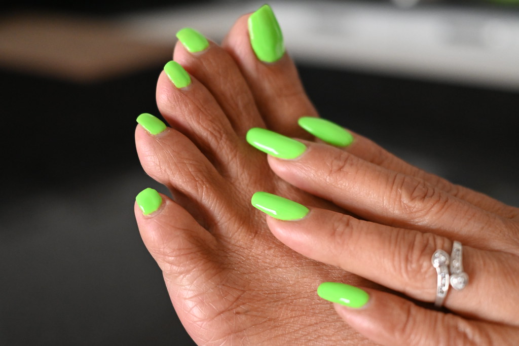A hand rests on a foot, both featuring vibrant neon green nail polish against a dark blurred background. — illustrating “Chanel Le Vernis 307 Orange Fizz” on Florence and Mary