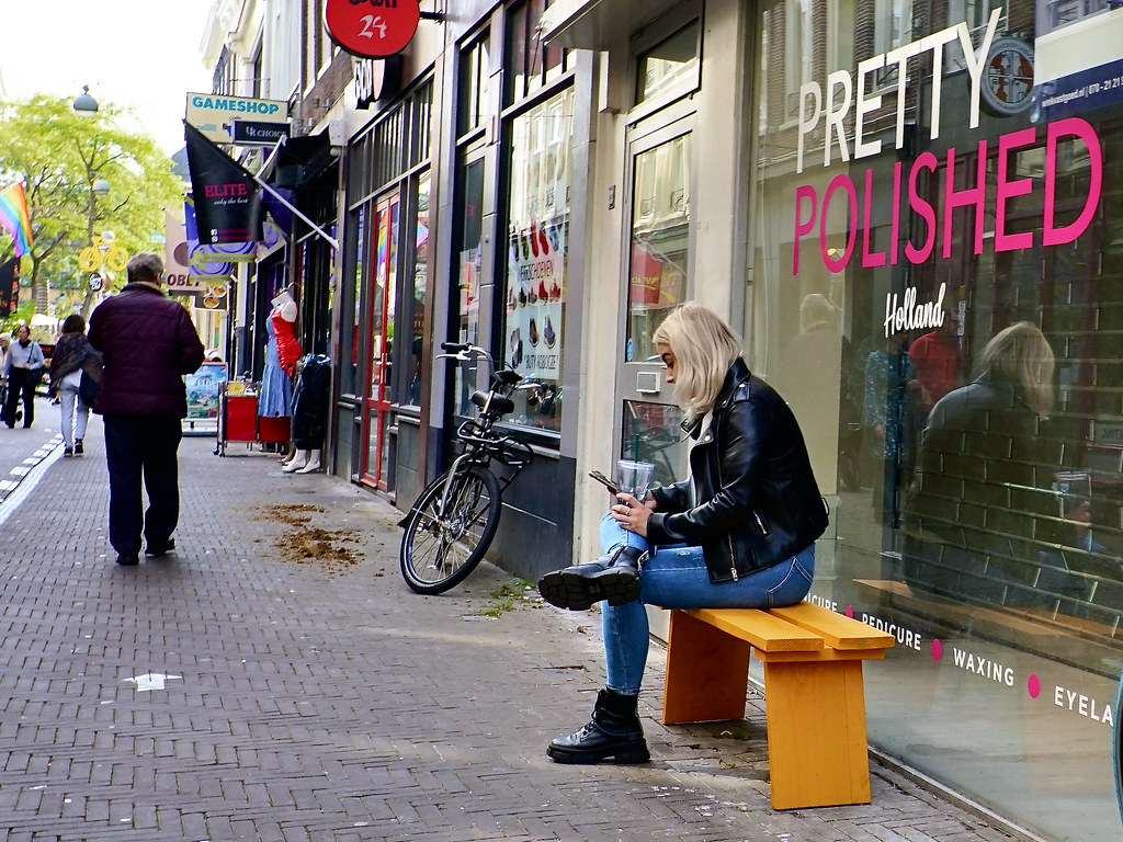 A blonde woman in a black leather jacket sits on a yellow bench outside a pink-lettered shop window. — illustrating “Chanel Le Vernis Nail Colour - 508 Pirate” on Florence and Mary