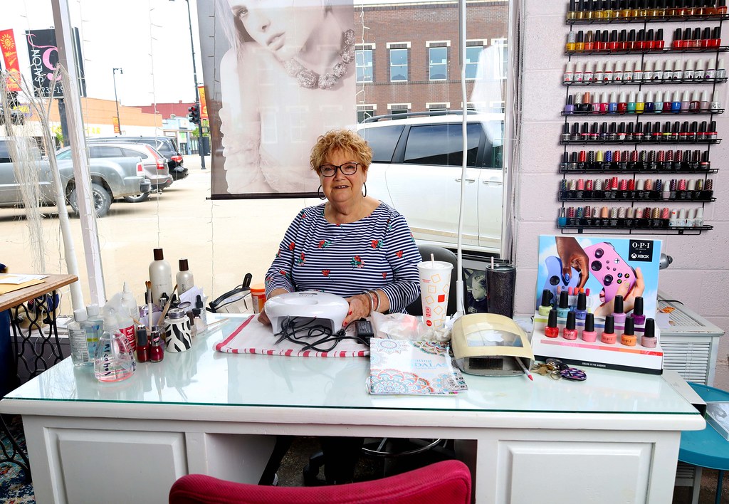 A smiling woman in a striped shirt sits at a white manicure desk stocked with colorful polish bottles. — illustrating “Chanel Le Vernis Nail Colour: 657 Azure” on Florence and Mary