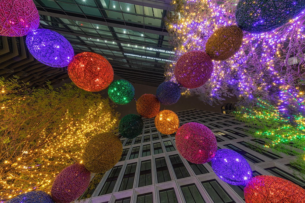 Colorful woven lanterns and fairy-lit trees hang between tall city buildings in an upward night view. — illustrating “Christmas Saving Tip | Look after the pennies” on Florence and Mary