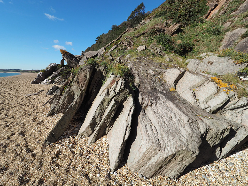 Sharply tilted grey rock slabs cover a pebble beach beside a green hillside under a cloudless blue sky. — illustrating “Clacton on Sea Day trip” on Florence and Mary