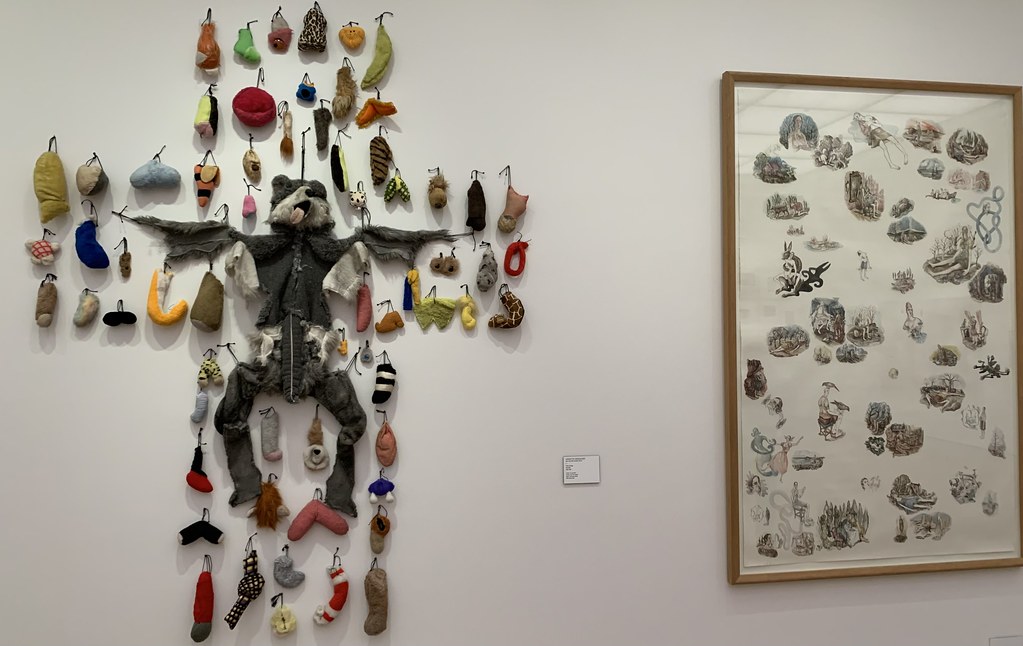 A gallery wall shows a splayed gray plush creature surrounded by colorful hanging objects beside a framed drawing. — illustrating “Collection Work The Colour Nail Polish” on Florence and Mary