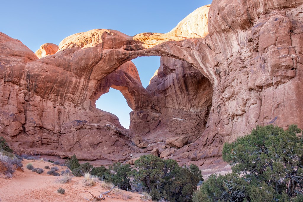 A massive reddish-orange sandstone double arch stands against a clear blue sky with green desert shrubs below. — illustrating “December Empties” on Florence and Mary