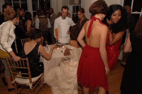 Guests in formal attire including white and red dresses gather around a candlelit table at a crowded event. — illustrating “December Empties” on Florence and Mary