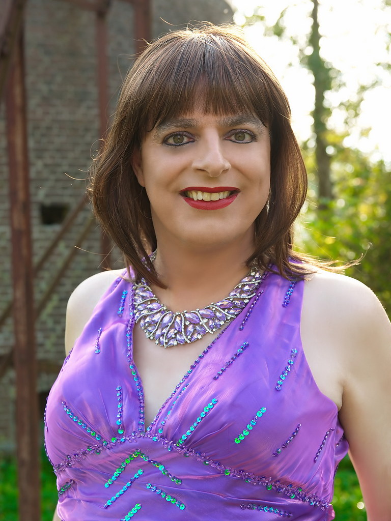 A person with brown hair and bangs smiles outdoors in a sequined purple dress and matching statement necklace. — illustrating “Desk to Disco | Lovely Lashes” on Florence and Mary