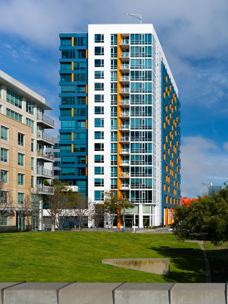 A tall white apartment building with blue and orange accents rises behind a grassy park under blue sky. — illustrating “Dining out on a diet - Pizza Express” on Florence and Mary