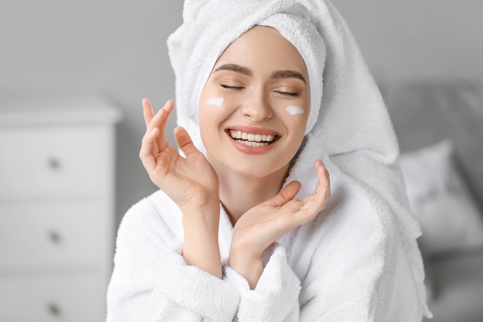A laughing woman in a white bathrobe and towel turban has cream dabs on her cheeks in a bright room. — illustrating “Facial Oil Favourites” on Florence and Mary