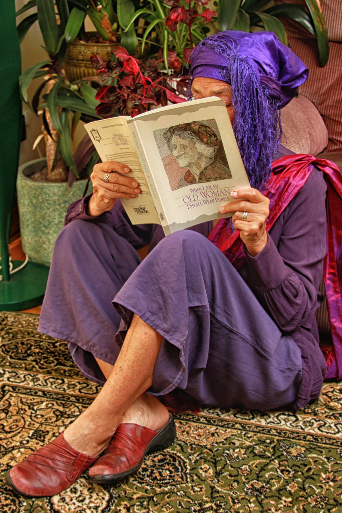 An older woman in vibrant purple clothing and a fringed headscarf reads a book on a patterned rug by plants. — illustrating “Fat Friends!” on Florence and Mary