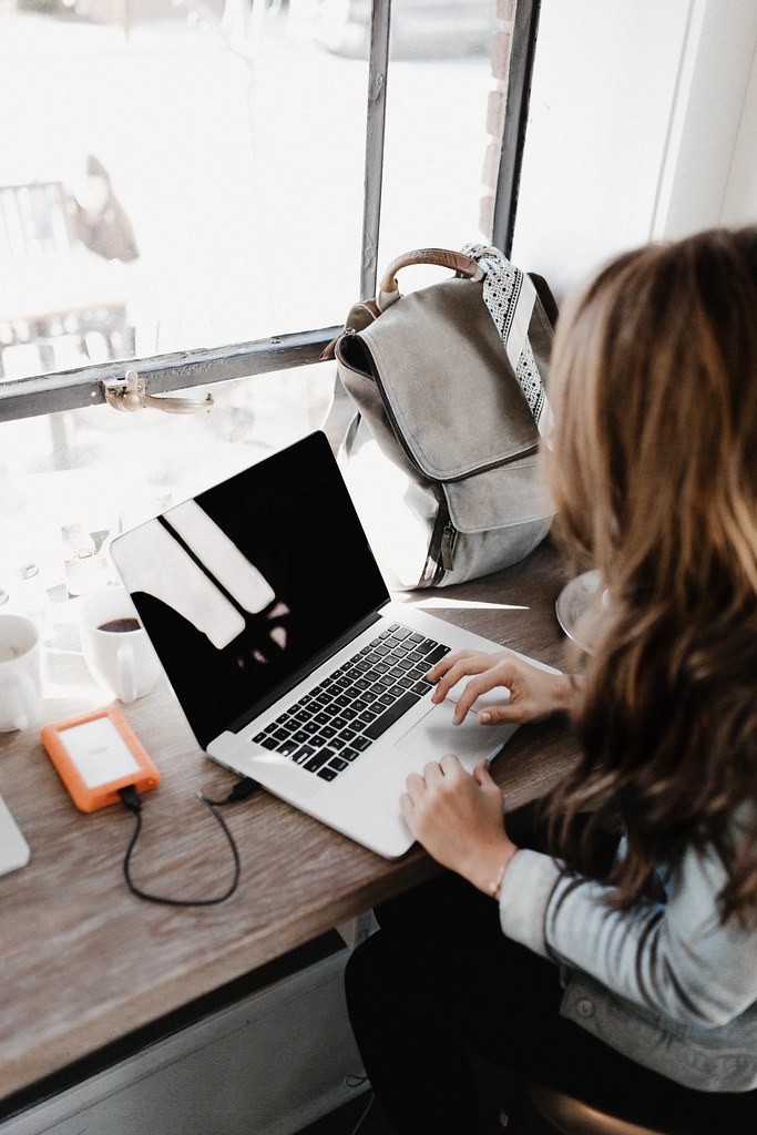 A person with long wavy brown hair works on a silver laptop beside a window with an orange hard drive. — illustrating “From Desk to Disco | Hair Heroes” on Florence and Mary
