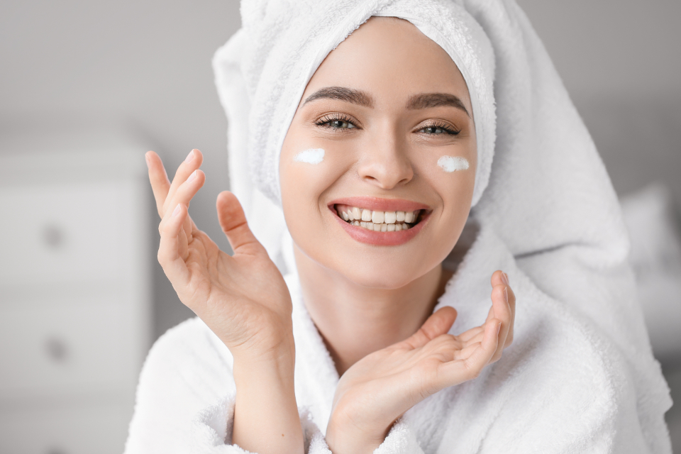 A smiling woman in a white bathrobe and towel turban shows cream dabs on her cheeks with hands raised. — illustrating “Graham Skincare - now available in the UK” on Florence and Mary