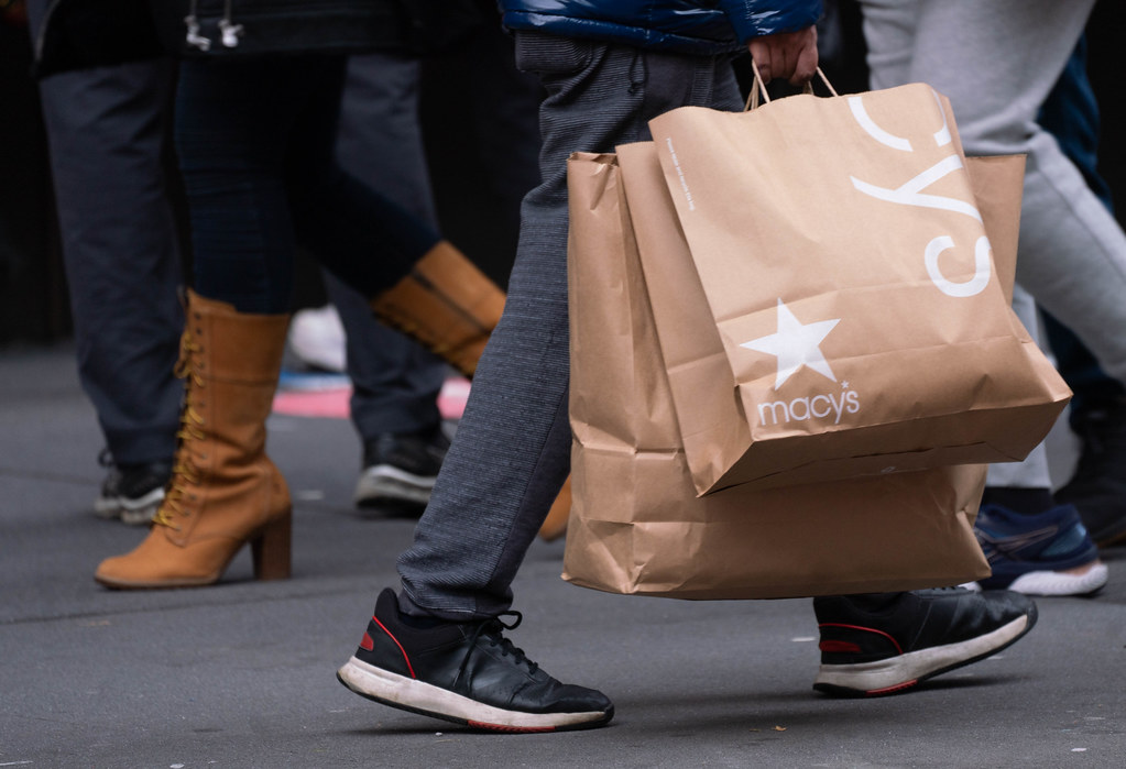 A person in grey trousers carries large brown Macy paper shopping bags while walking a crowded sidewalk. — illustrating “Holiday Essentials Haul” on Florence and Mary