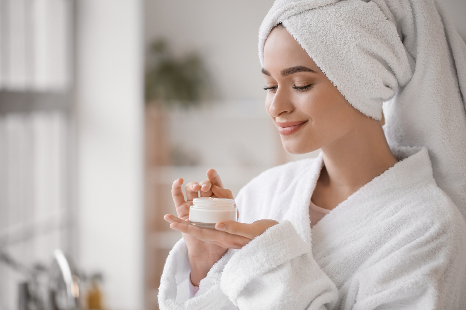 A smiling woman in a white bathrobe and towel wrap holds an open jar of white cream in a bright room. — illustrating “Holiday Skincare Regime” on Florence and Mary