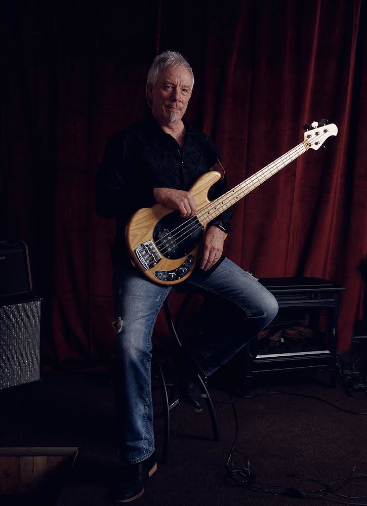 A middle-aged man with grey hair sits on a stool holding a light wood electric bass before red curtains. — illustrating “In Flight Entertainment // Movies” on Florence and Mary