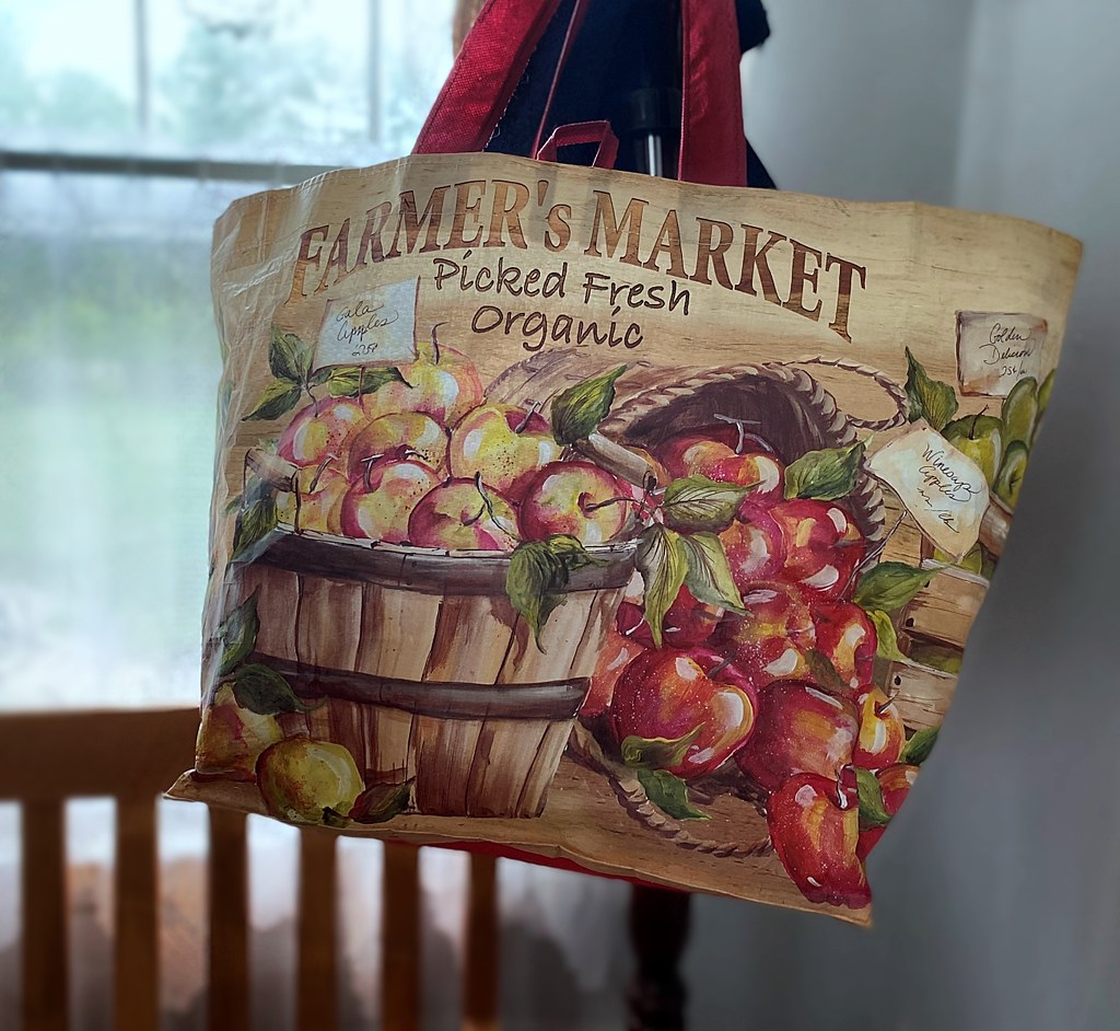 A tan reusable bag with red handles shows a rustic illustration of red and yellow apples in baskets. — illustrating “In Flight Essentials | Short Haul Flights” on Florence and Mary