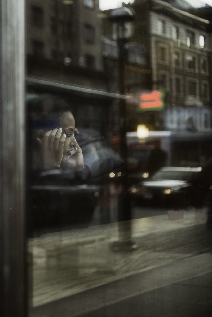 A young woman seen through a reflective window talks on a smartphone with blurred city lights behind. — illustrating “The Lost Passport Saga - and tips if you lose yours!” on Florence and Mary