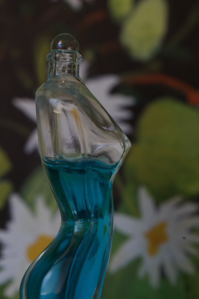 A clear torso-shaped perfume bottle filled with bright blue liquid sits before a soft floral backdrop. — illustrating “Jil Sander No 4 | A Beauty Enigma” on Florence and Mary