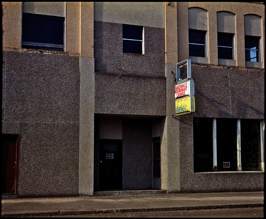 A grey pebbled building facade shows dark windows and a vintage Oakesdale Restaurant Lounge hotel apartments sign. — illustrating “June Empties | Beauty” on Florence and Mary