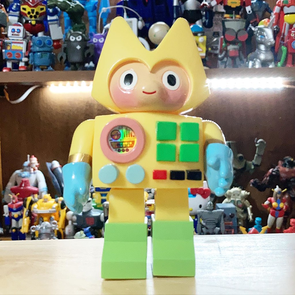 A yellow cat-eared toy robot with blue hands and green feet stands before a shelf of other figures. — illustrating “June Empties” on Florence and Mary