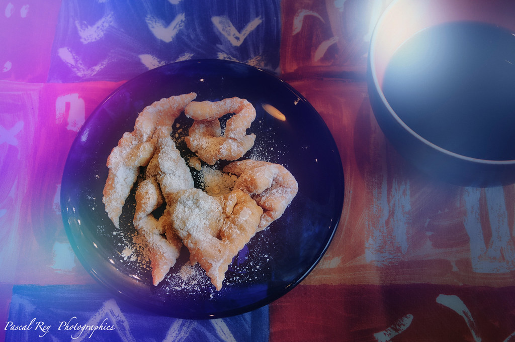 Golden fried pastries dusted with powdered sugar sit beside a cup on a patterned tablecloth with purple flares. — illustrating “Laduree Limited Edition Sonny Angel Figure” on Florence and Mary