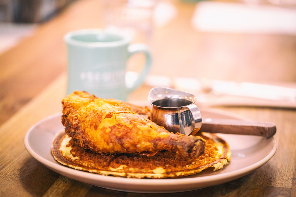 Crispy golden fried chicken rests on a thin round waffle with a metal syrup pitcher on a wooden table. — illustrating “Le Larry at Belgique” on Florence and Mary