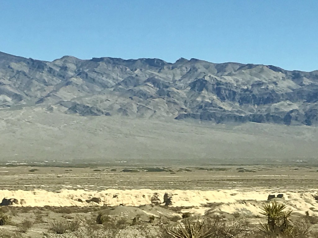 A wide desert landscape of light rocky terrain and sparse shrubs leads to rugged grey mountains under blue sky. — illustrating “Leaving Las Vegas” on Florence and Mary