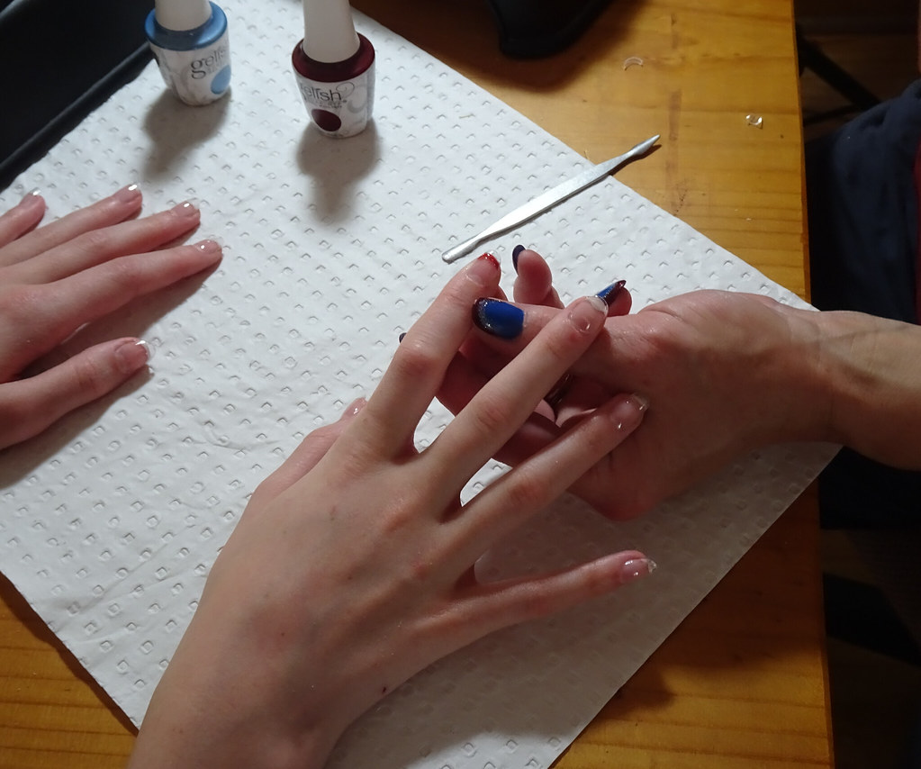 Hands receive a manicure on a white towel with blue and burgundy polish bottles on a wooden table. — illustrating “Leighton Denny Crystal Nail File | An Update” on Florence and Mary