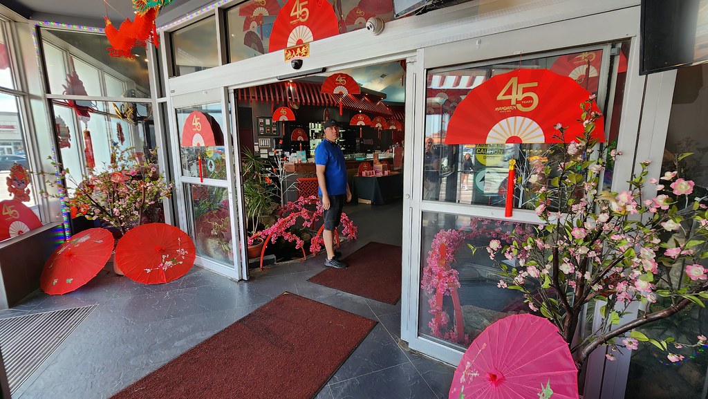 A man in a blue shirt stands at an entrance decorated with red commemorative fans, cherry blossoms, and parasols. — illustrating “London Burger War // Shake Shack Vs Five Guys” on Florence and Mary