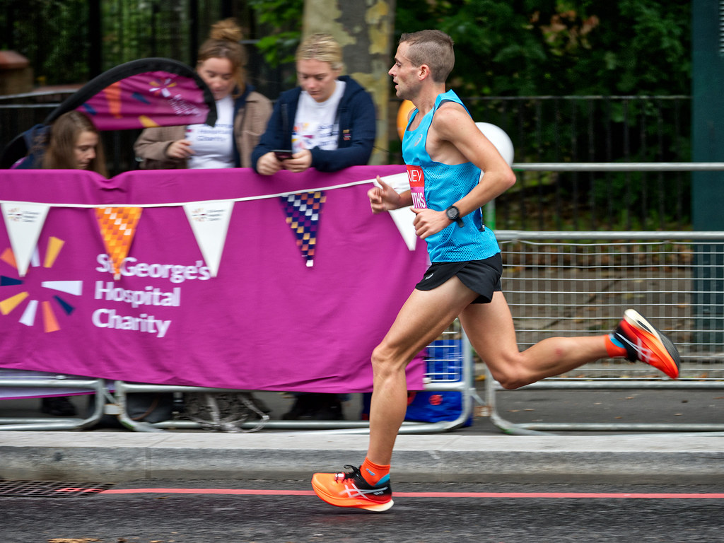 A male runner in a bright blue tank top and orange shoes sprints past a purple charity banner. — illustrating “Marathon Man” on Florence and Mary