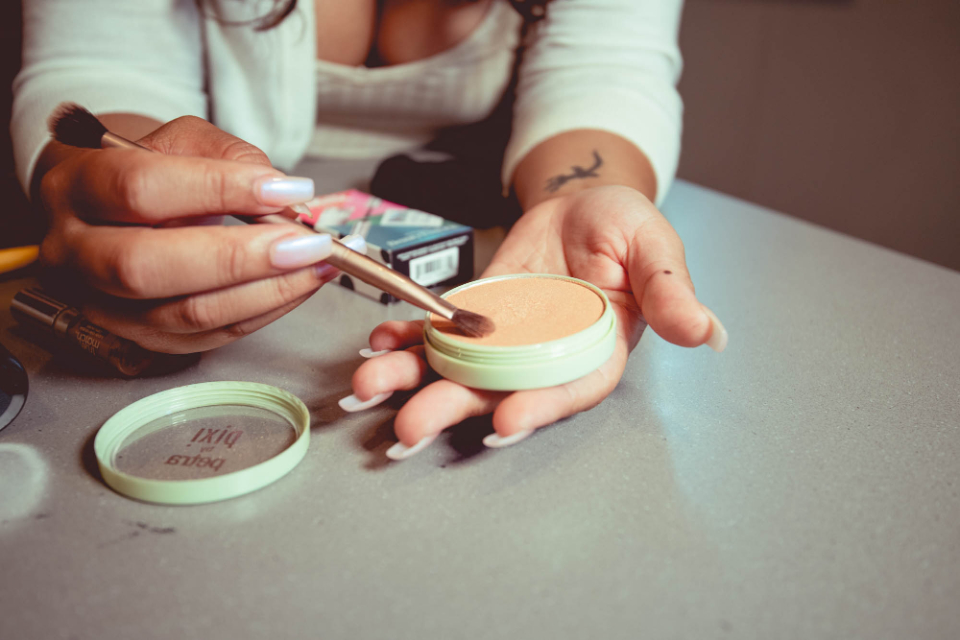 Hands with iridescent nails and a bird tattoo dip a brush into a pale green compact of peach powder. — illustrating “May Empties” on Florence and Mary