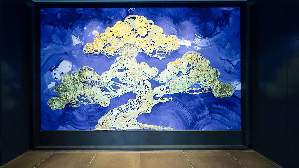 An intricate gold mechanical bonsai tree sculpture stands against a swirling vibrant blue painted gallery wall. — illustrating “Maybelline Master Glaze Cream Blush” on Florence and Mary