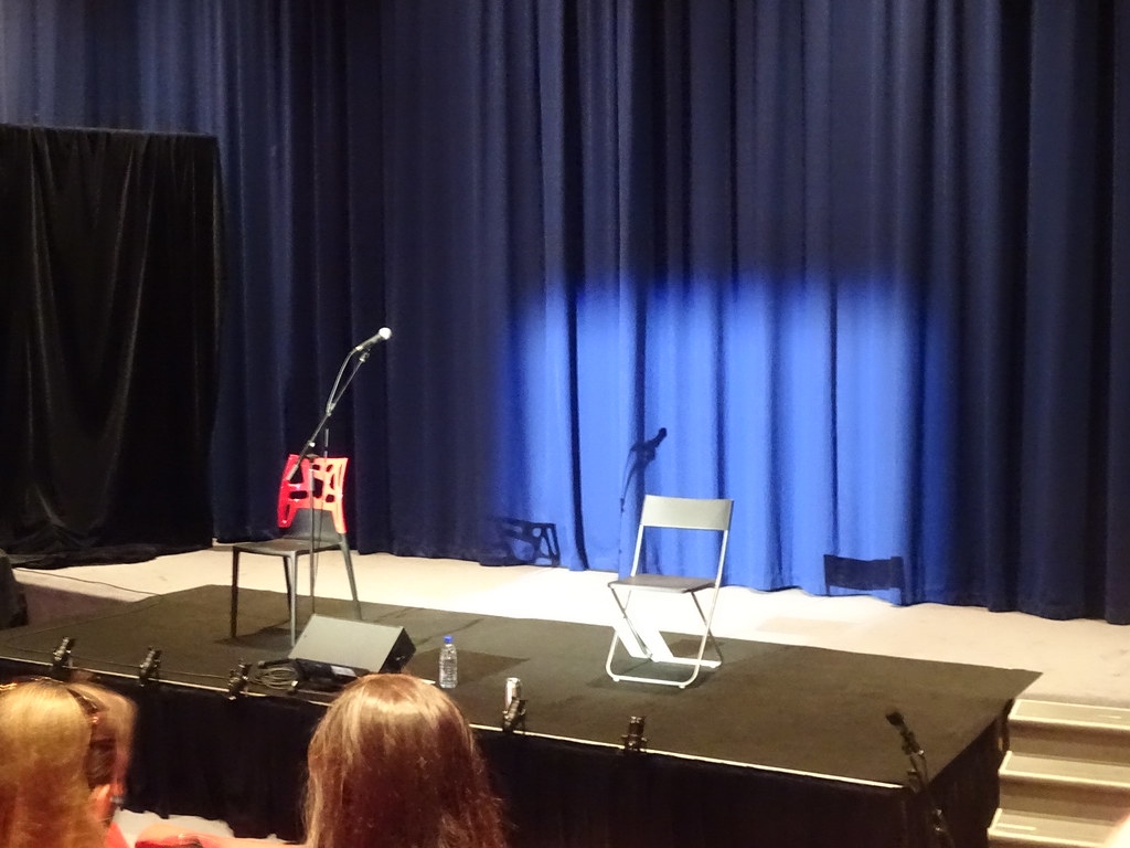 An empty stage with two chairs and a microphone sits before navy curtains under a spotlight. — illustrating “Mickey Flanagan - Shall we go out out?” on Florence and Mary