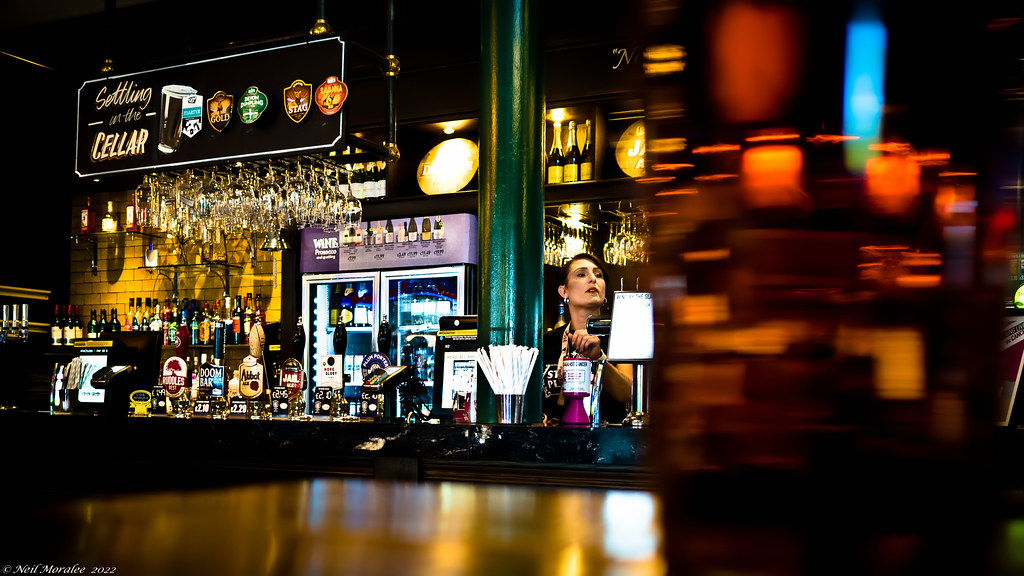 A female bartender works behind a dark pub counter with beer taps, hanging glasses, and a green pillar. — illustrating “Moosehead and Milk” on Florence and Mary