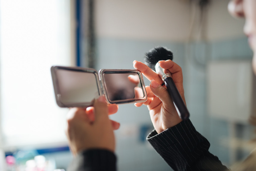 A person in a dark pinstriped sleeve holds a silver compact mirror and black makeup brush before a window. — illustrating “My Morning Skincare Regime” on Florence and Mary