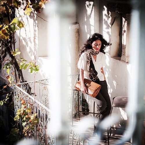 A woman in a white blouse and patterned scarf holds a brown leather clutch on a vine-covered balcony. — illustrating “#mydubai” on Florence and Mary