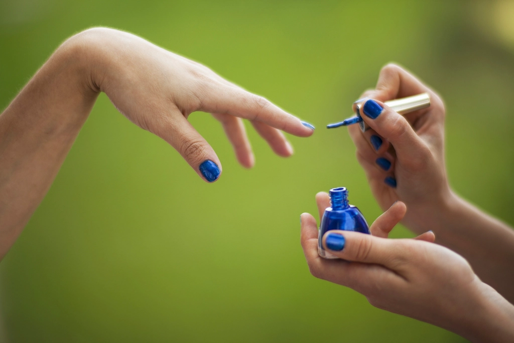 Two hands apply vibrant royal blue nail polish outdoors against a soft creamy green blurred background. — illustrating “Nailbox Subscription Service” on Florence and Mary