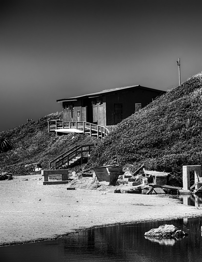 Black-and-white hillside building above a sandy beach beside a still reflective pool of water. — illustrating “Not So Shabby” on Florence and Mary
