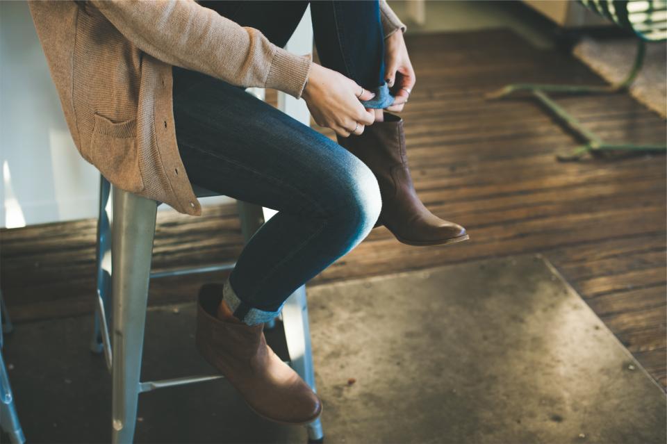 Person in beige cardigan and cuffed jeans sits on a silver stool adjusting brown ankle boots. — illustrating “OOTD: Office Chic” on Florence and Mary